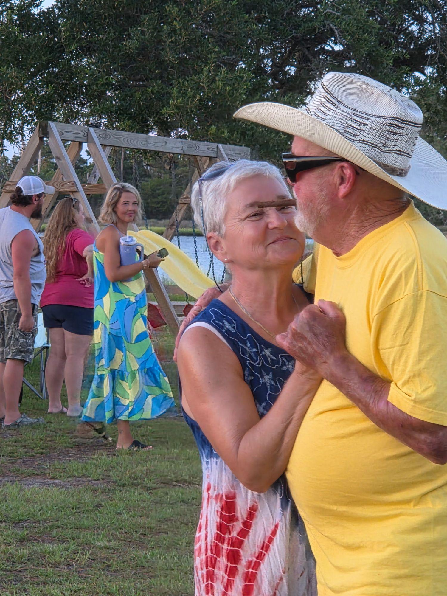 A couple dancing on the lawn by the swing set, she in a red, white and blue dress and he in a straw cowboy hat.
