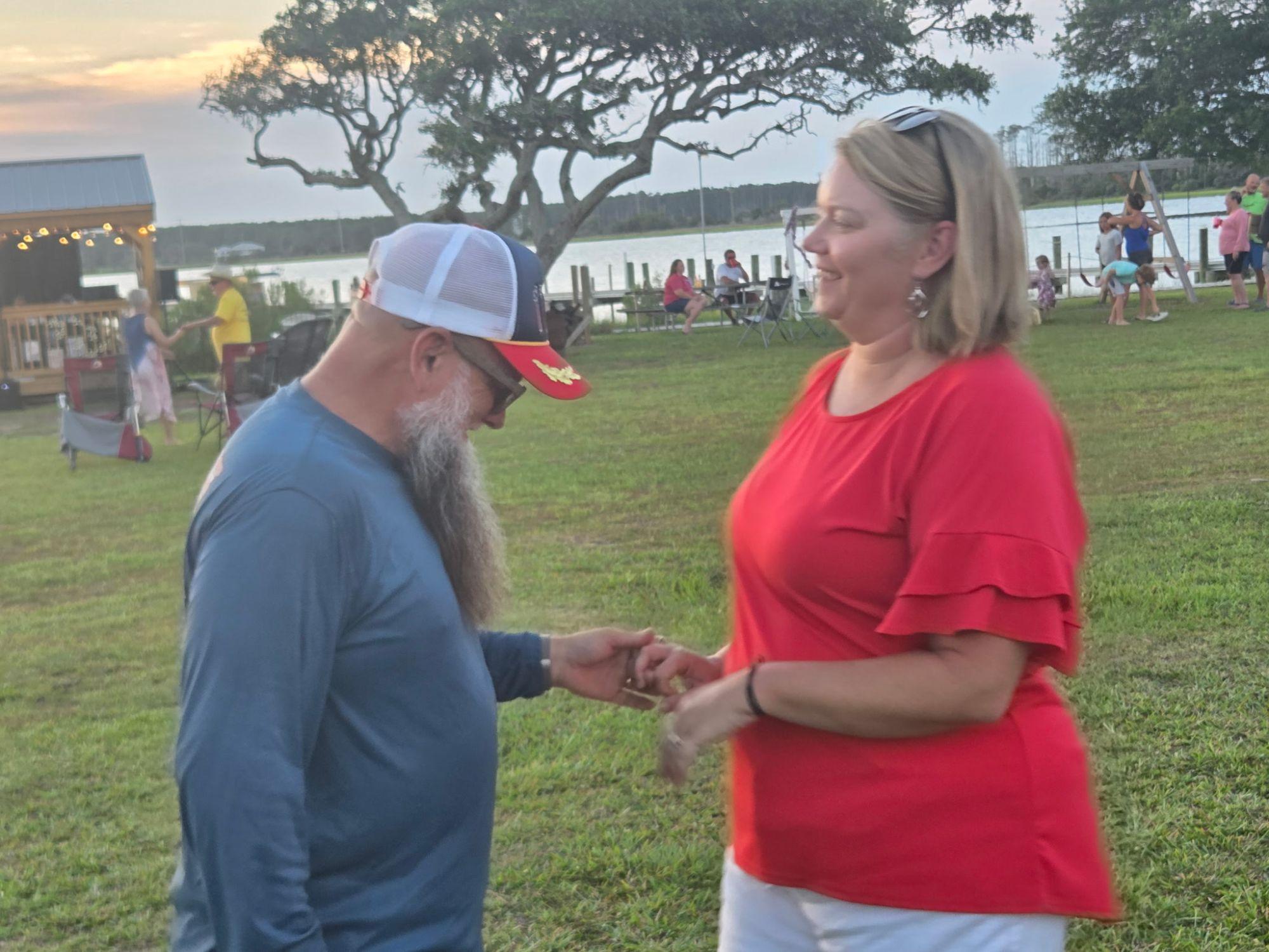 A man in a trucker hat and a woman in a red top dancing on the grass, with the pavilion, the creek and other guests behind them.