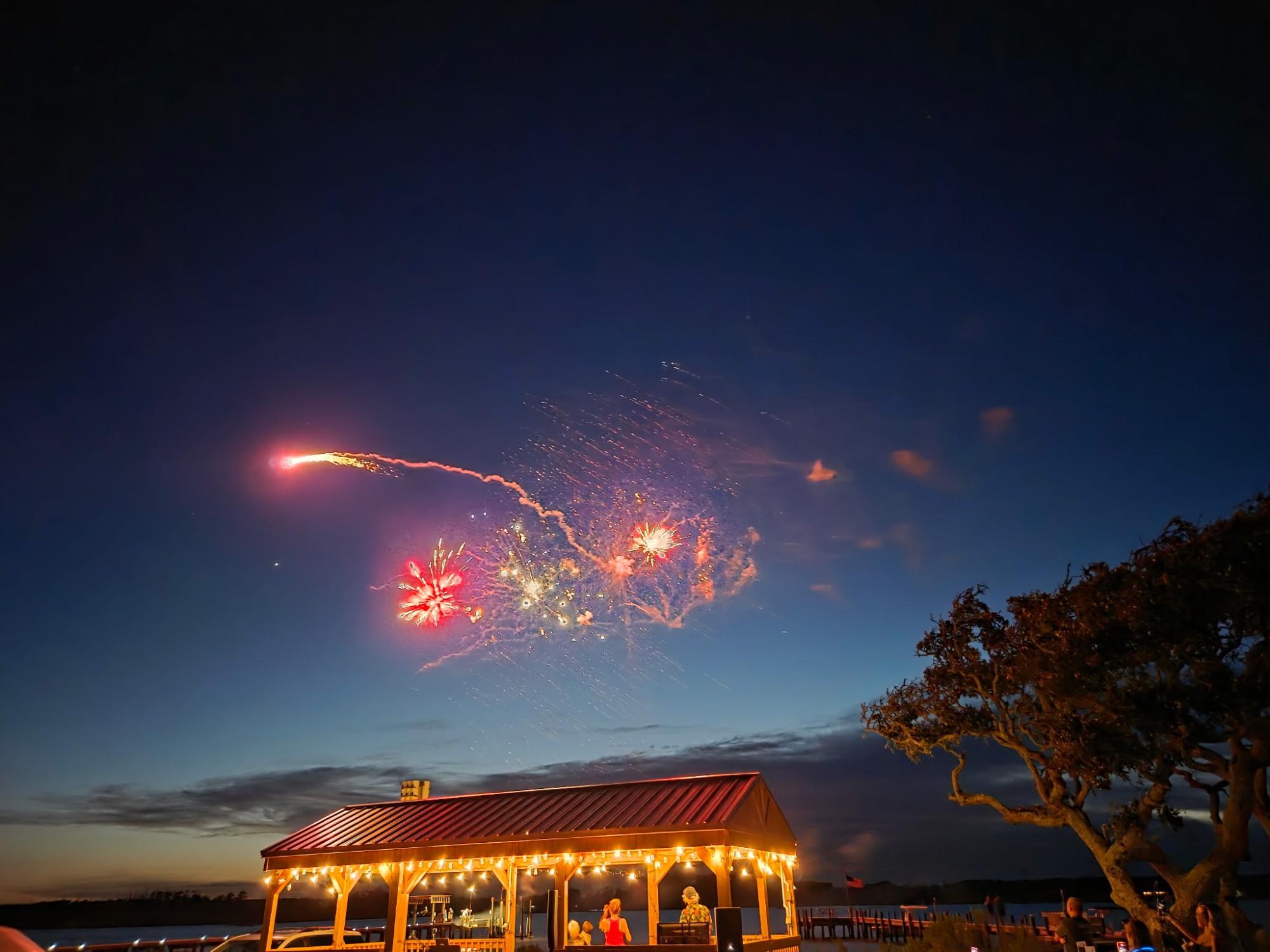 Fireworks in the night sky over the pavilion by the creek, its roofline strung with lights and a few people gathered underneath.