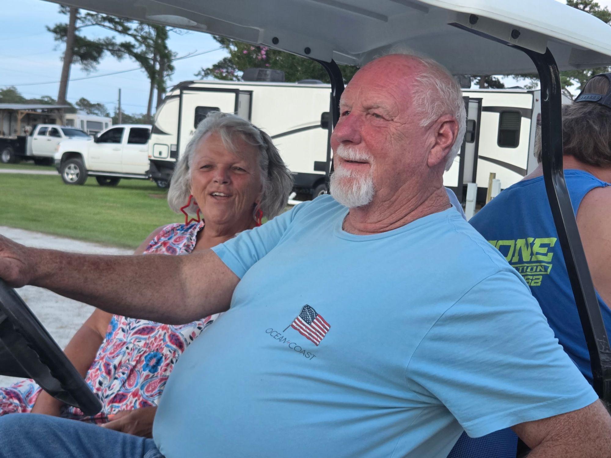 A couple riding in a golf cart past the RV sites, she wearing red star earrings and he in a flag T-shirt.