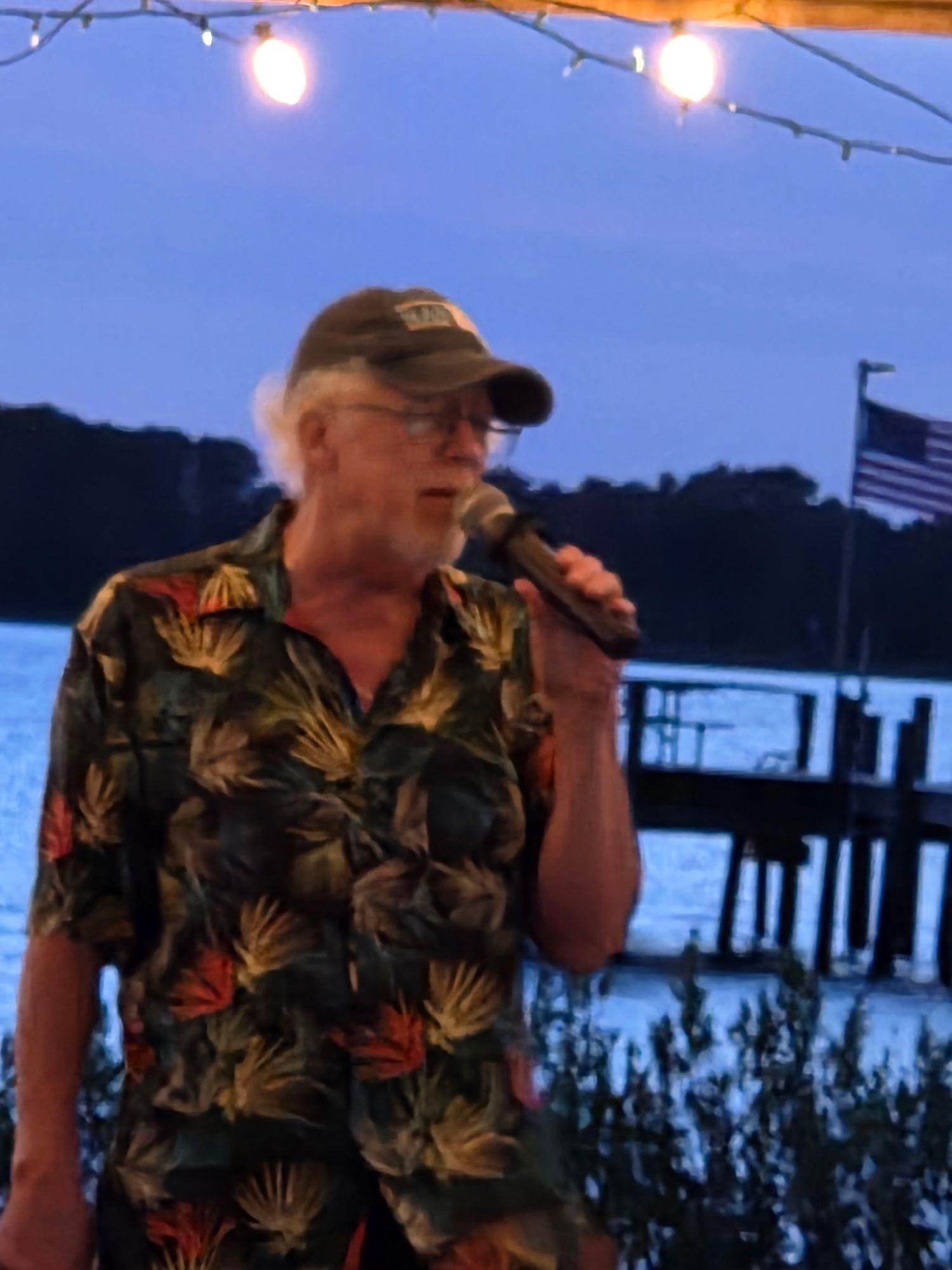 A man in a ball cap and a palm-print shirt singing into a microphone under string lights, with the pier and an American flag behind him.