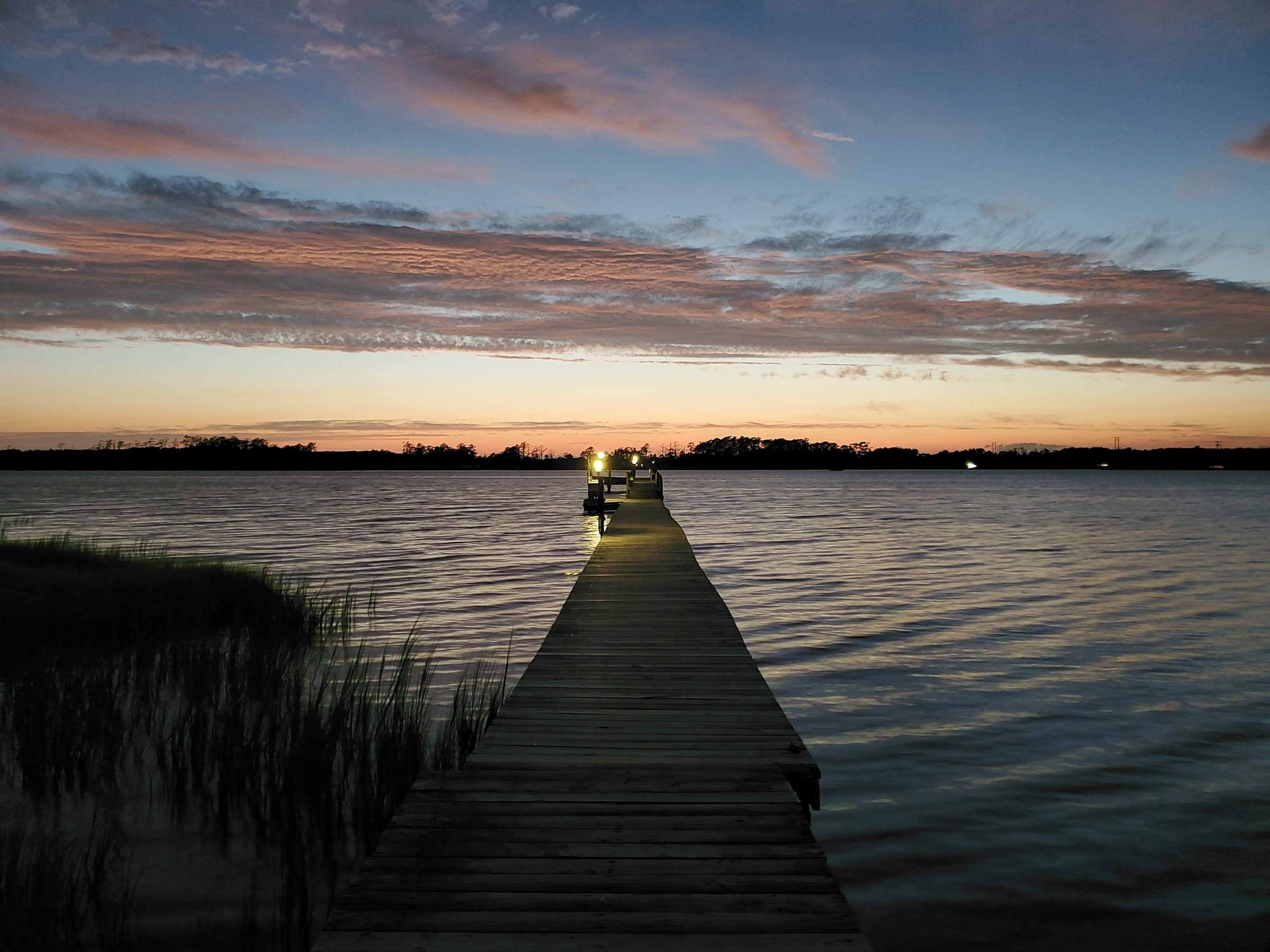 A long wooden pier running out over a creek at sundown, with marsh grass in the foreground and a pink and gray sky.